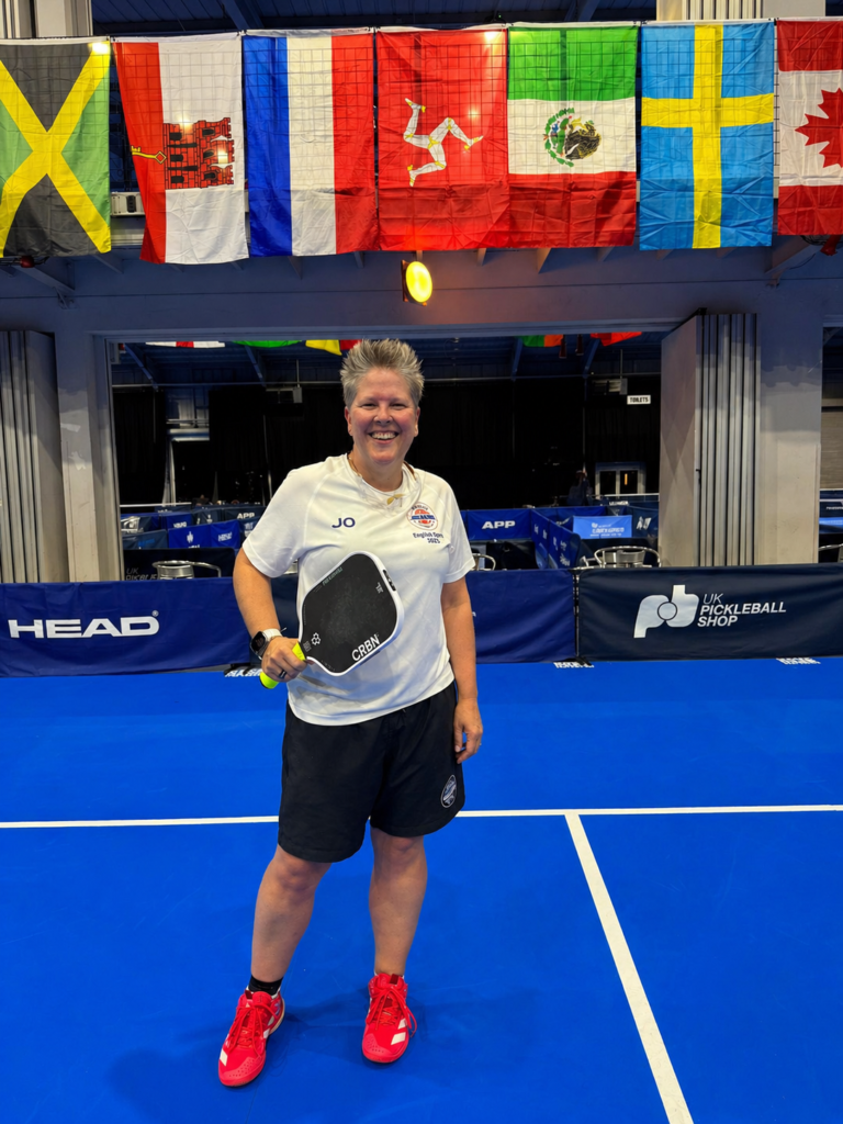 Smiling woman in white t‑shirt and black shorts stands on a blue pickleball court, holding a paddle, with colorful flags overhead.