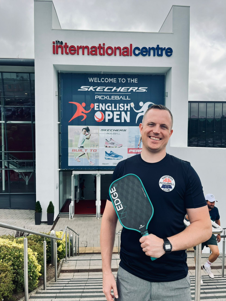 Smiling man holding a pickleball paddle in front of a banner for the English Open at The International Centre.