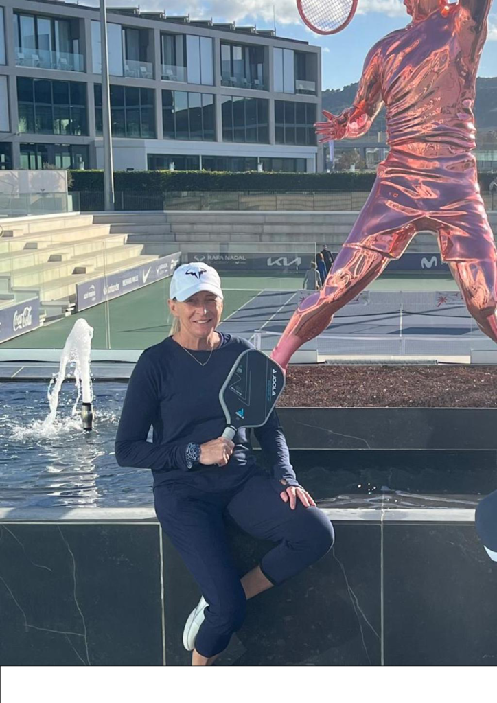Smiling woman in dark athletic wear and a white cap sits by a fountain, holding a paddle-style tennis racquet, with a large pink tennis-player statue and an outdoor court behind her.